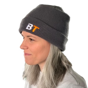 May include: A gray beanie with a folded brim. The beanie features the embroidered letters "BT" in white and orange. The hat is made of a ribbed knit material and is worn by a person.