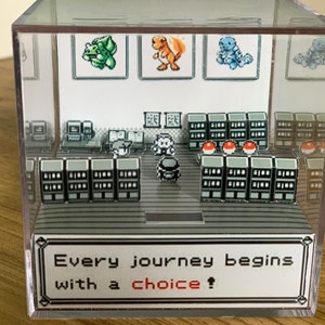 May include: A clear acrylic cube displaying a pixelated video game scene. The scene includes characters, shelves, and the text "Every journey begins with a choice!" in a white frame. The cube is set against a wooden background.