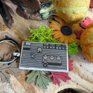 May include: A wooden keychain featuring a pixelated image of a video game character. The keychain is attached to a silver key ring. The background of the image is grey and the character is brown.