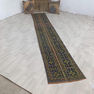 Op de afbeelding: Een lange, smalle loper met een herhalend geometrisch patroon in tinten donkerblauw, groen en oranje. Het kleed heeft een gefranjerde rand en wordt getoond naast twee vierkante sierkussens met vergelijkbare patronen en een houten kist.
