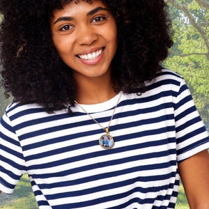 May include: A gold-colored necklace with a round pendant featuring a colorful image. The necklace is worn over a navy and white striped t-shirt. The background is a blurred outdoor scene with green foliage.