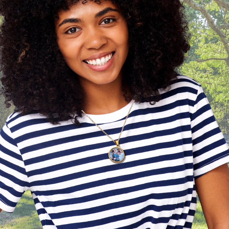 May include: A gold-colored necklace with a round pendant featuring a colorful image. The necklace is worn over a navy and white striped t-shirt. The background is a blurred outdoor scene with green foliage.