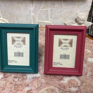 May include: Two rectangular picture frames, one teal and one magenta, each with a white mat and the text "MOMENTS" and "2 1/2" x 3 1/2"" printed on the mat. The frames are displayed on a marble-patterned surface.