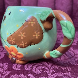 May include: A green ceramic mug with a brown owl design. The owl has a brown body with orange flowers and yellow dots. The mug has a brown handle with green leaves.