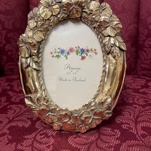 May include: Gold oval picture frame with a floral design. The frame is 3.5 inches by 5 inches and says "Amora Made in Thailand".