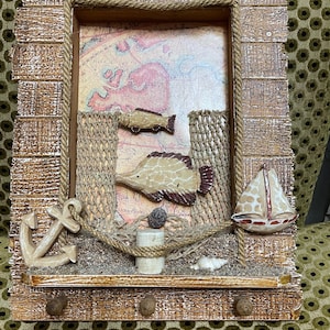 May include: A decorative wooden wall hanging with a nautical theme. It features a framed design with a map background, two fish, an anchor, a sailboat, and a rope net. The frame has three small hooks at the bottom.
