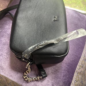 May include: Black rectangular crossbody bag with a silver chain strap and zipper. The bag has a small silver logo on the front. The bag is made of a textured material and has a long strap.
