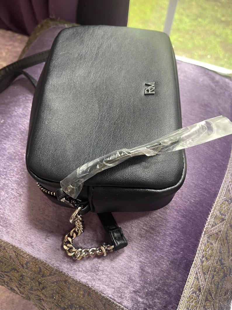 May include: Black rectangular crossbody bag with a silver chain strap and zipper. The bag has a small silver logo on the front. The bag is made of a textured material and has a long strap.