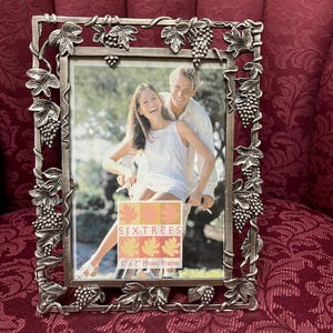 May include: Silver-toned metal photo frame with a grape vine design. The frame holds a 5" x 7" photo and features the text "SIXTREES". The frame is displayed on a patterned red surface.