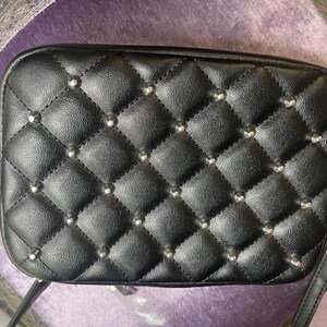 May include: Black rectangular crossbody bag with a quilted pattern and silver-colored studs. The bag has a shoulder strap and a tag attached. The bag is made of a leather-like material.