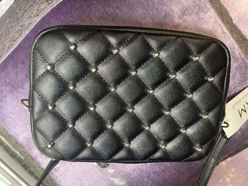 May include: Black rectangular crossbody bag with a quilted pattern and silver-colored studs. The bag has a shoulder strap and a tag attached. The bag is made of a leather-like material.
