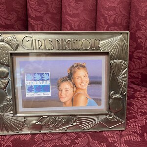 May include: Silver-toned picture frame with a raised inscription "Girls Night Out" and a smaller inscription "Sixtrees 4" x 6" Photo Frame". The frame features a design of palm leaves, cocktails, and umbrellas. The frame has a photo of two women smiling at the camera.