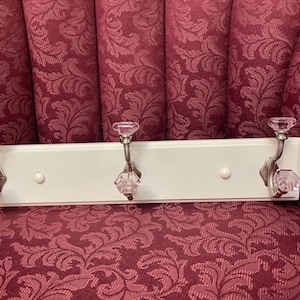 May include: White wooden coat rack with three hooks. Each hook has a silver finish and a clear glass knob with a pink tint.