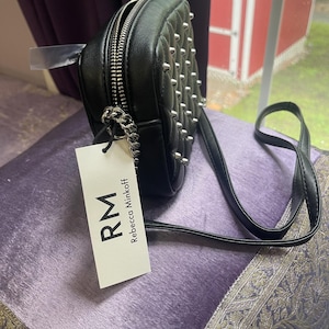 May include: Black, studded crossbody bag with a silver zipper and chain detail. The bag has a long, thin strap and a tag that reads "RM Rebecca Minkoff". The bag is sitting on a purple surface.