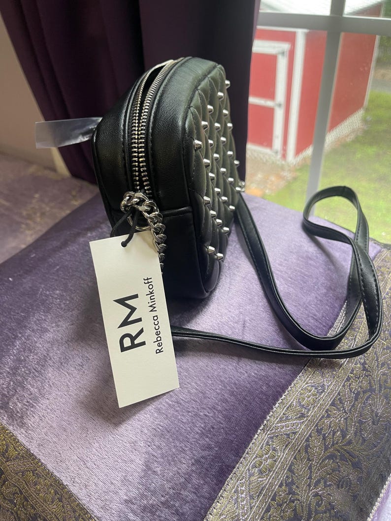 May include: Black, studded crossbody bag with a silver zipper and chain detail. The bag has a long, thin strap and a tag that reads "RM Rebecca Minkoff". The bag is sitting on a purple surface.