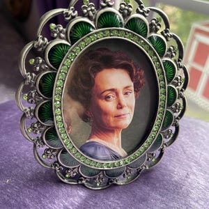 May include: Oval silver-toned picture frame with a portrait of a woman. The frame has a scalloped edge with green enamel and green rhinestone accents. The frame is sitting on a purple surface.