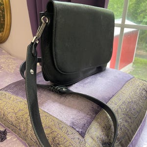 May include: A black leather crossbody bag with a flap closure and adjustable strap. The bag is resting on a purple pillow with gold trim. The bag has silver-tone hardware.