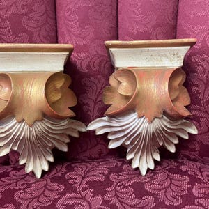 May include: Two ornate, decorative wall brackets made of wood with a white and gold paint finish. The brackets have a scalloped design and a flat top.
