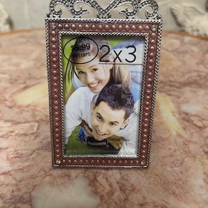 May include: A rectangular photo frame with a decorative silver top in a heart shape. The frame is 2x3 inches and has a pinkish-brown border with small, round embellishments. The frame holds a photo of a smiling couple.