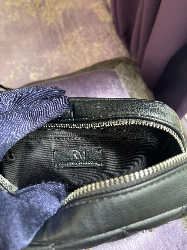 May include: Black leather crossbody bag with a silver zipper and studded accents. The interior features a black lining and a label that reads "RM REBECCA MINKOFF". The bag is open, revealing the inside.