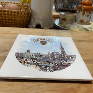 May include: Square ceramic tile featuring a detailed illustration of Vienna's cityscape, including prominent buildings and a church spire. The tile has a white border and the word "Wien" is written across the top.