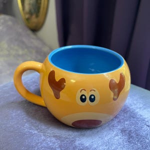 May include: A whimsical, round ceramic mug with a yellow exterior featuring a cartoon reindeer face. The mug has a blue interior and a yellow handle. The reindeer design includes brown antlers, eyes, and a red nose.