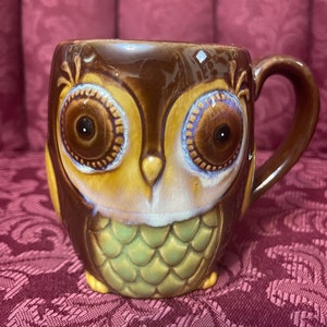 May include: A brown ceramic owl mug with large brown eyes and a green and brown owl design. The mug has a handle on the right side.