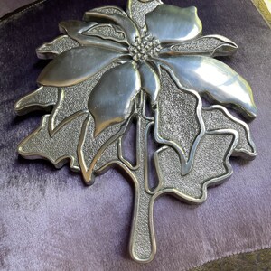 May include: A silver metal poinsettia ornament with a textured surface. The ornament is shaped like a poinsettia flower with leaves.
