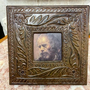 May include: Square, ornate picture frame with a portrait of a man. The frame is a dark, bronzed color with floral and leaf detailing. The portrait is a close-up of a man's face, with a focus on his eyes and beard.