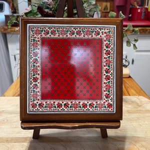 May include: A red and green ceramic tile with a floral pattern is displayed on a wooden easel. The tile has a red center with a white and green floral border.