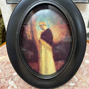 May include: An oval, black framed artwork featuring a painting of a person in a long, light-colored garment. The painting has a warm color palette with gold, brown, and purple hues. The frame has a beaded edge.