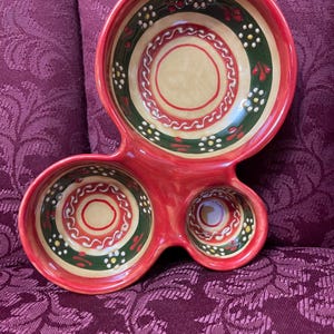 May include: A red ceramic serving dish with three connected bowls. The bowls have a beige interior with red and green decorative rings and white and yellow accents. The dish is on a patterned purple surface.