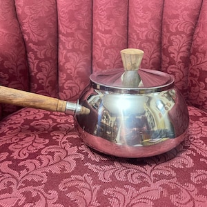 May include: A shiny, stainless steel saucepan with a wooden handle and lid. The handle and lid knob are made of light-colored wood. The pot sits on a red patterned fabric.