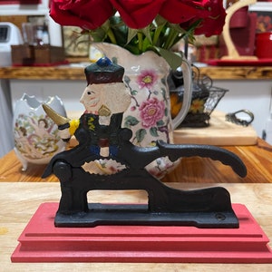 May include: A black cast iron nutcracker shaped like a soldier with a red and blue uniform, holding a nut in his hand. The nutcracker is on a red wooden base.