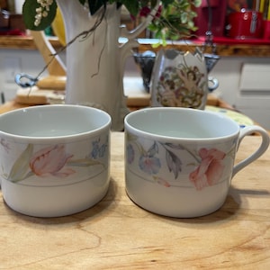 以下が含まれることがあります： 花柄の白いセラミック製のコーヒーカップが2つ。1つのカップにはピンクとブルーの花、もう1つのカップにはピンクとパープルの花が描かれています。