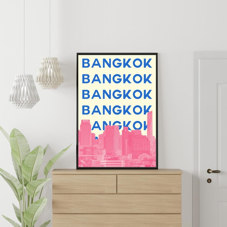 Puede incluir: Un cuadro enmarcado con la palabra "BANGKOK" en azul, repetida cinco veces, sobre un fondo crema. Debajo del texto hay una silueta rosa de la ciudad. La obra de arte se exhibe sobre una c&oacute;moda de madera.