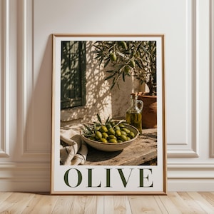 Könnte beinhalten: Ein gerahmter Kunstdruck mit einer Schale voller grüner Oliven, einer Glasflasche mit Olivenöl und einem Olivenzweig. Die Szene ist auf einer Holzoberfläche mit einem strukturierten Tuch arrangiert. Das Wort OLIVE steht in großen, dunkelgrünen Großbuchstaben darunter.