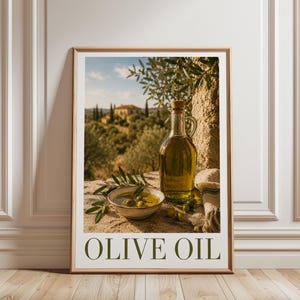 Op de afbeelding: Een ingelijste verticale wandkunstprint met een glazen karaf gevuld met goudkleurige olijfolie naast een kleine keramische kom met olijven op een stenen ondergrond, met een mediterraan landschap op de achtergrond en de tekst OLIVE OIL onderaan.