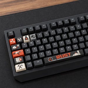 May include: A black mechanical keyboard featuring custom keycaps with icons and the text RUST. The keys include symbols of a radiation sign, a campfire, a gear, a rock, a rifle, a building, and a hammer and pickaxe on a dark background.