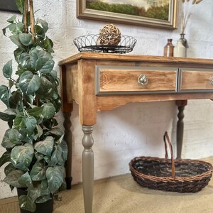 May include: A wooden console table featuring two drawers with metallic knobs, a natural wood grain top, and tapered legs painted in a muted grey tone with black feet. The table includes a wire basket on top and a woven wicker basket underneath.