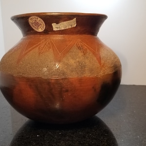 Puede incluir: Una olla de barro marrón con un diseño geométrico. La olla tiene una boca ancha y un cuerpo redondeado. Está decorada con un patrón de triángulos y líneas.