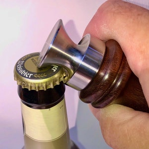 May include: A close-up of a wooden and stainless steel bottle opener opening a bottle of German Weissbier.