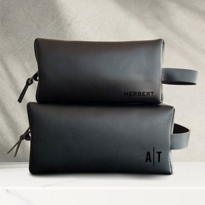 May include: Two black leather zippered pouches stacked on top of each other. The top pouch has the word "HERBERT" embossed on the front. The bottom pouch has the letters "A|T" embossed on the front.