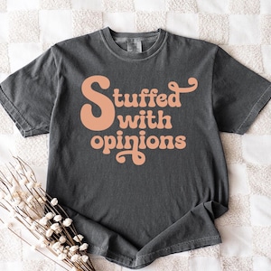 May include: Dark gray t-shirt with the phrase "Stuffed with opinions" in a retro, peach-colored font. The shirt is made of a soft material and has short sleeves. The shirt is displayed on a white and beige patterned surface.