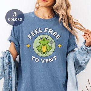 May include: A blue t-shirt with the text "FEEL FREE TO VENT" surrounding a cartoon frog. The frog is green and yellow, set inside a green circle. The shirt also has a graphic that says "5 COLORS".