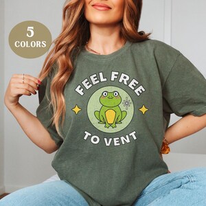 May include: Olive green t-shirt with a circular graphic of a cartoon frog and the text "FEEL FREE TO VENT". The shirt also has the text "5 COLORS" in a circle.