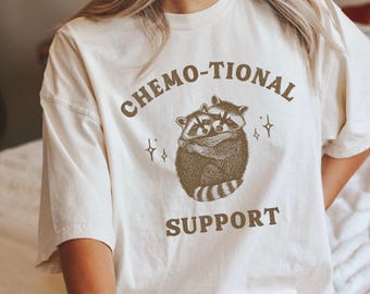 May include: An off-white short-sleeve t-shirt featuring a brown graphic of two raccoons hugging. The text CHEMO-TIONAL is printed in an arc above the raccoons, and the word SUPPORT is printed below them, accompanied by small star icons.