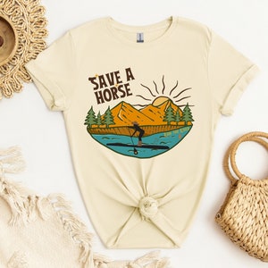 Puede incluir: Camiseta color crema con un gráfico de una persona haciendo paddle surf en un río, con montañas, árboles y un sol de fondo. El texto "Save a Horse Ride the River" se muestra encima de la escena. La camiseta está anudada a la cintura.