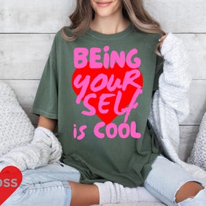 May include: A green t-shirt with the text "Being Your Self is Cool" in pink and red letters inside a pink heart.