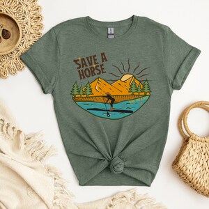 May include: Green t-shirt with a graphic of a person paddleboarding on a river, with mountains and trees in the background. The text "Save a Horse Ride the River" is above the image. The shirt is knotted at the waist.